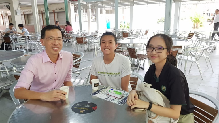 Three people sitting together at a metal table in an open-air cafe, smiling at the camera.